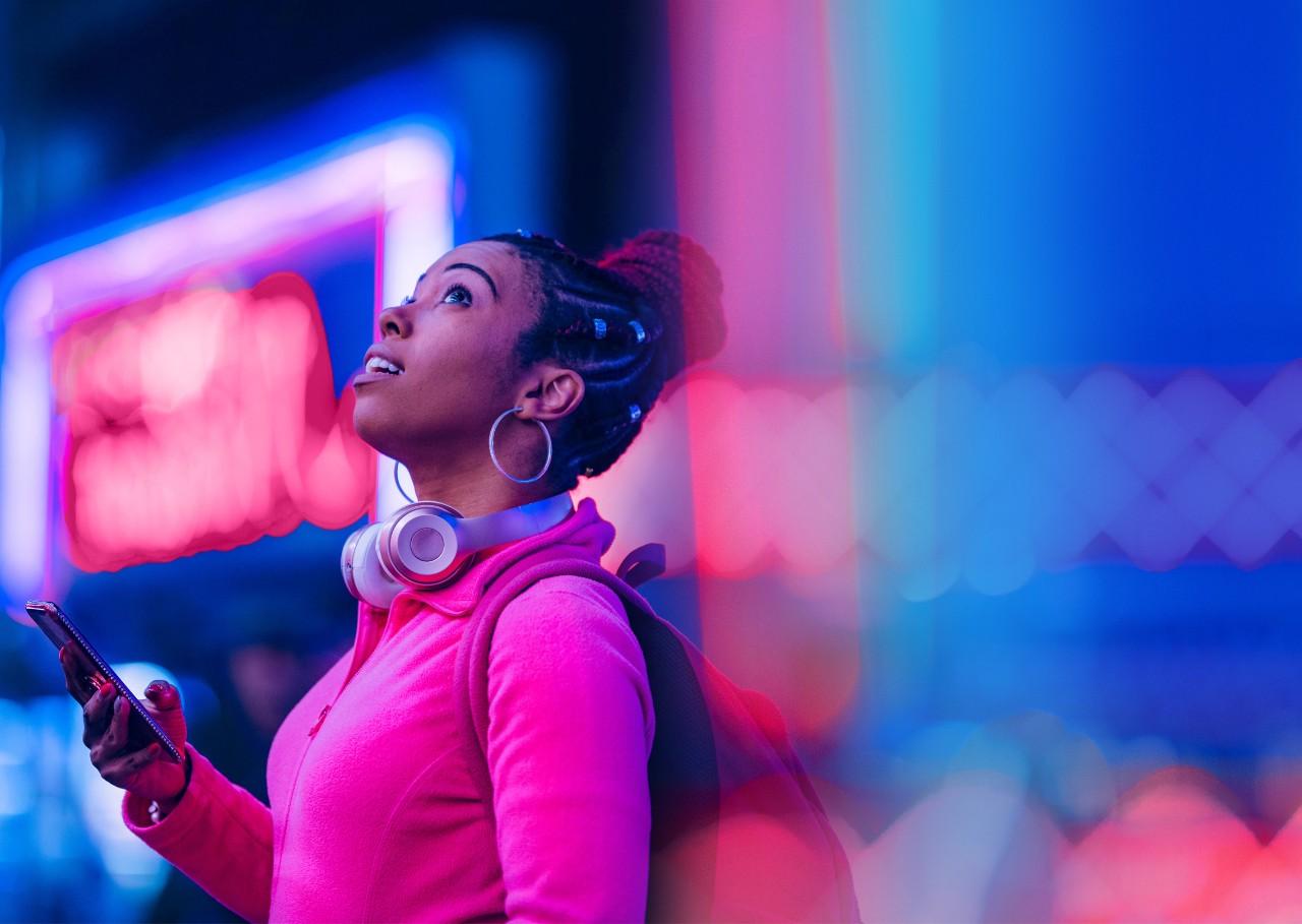 年轻的黑人女子穿着粉色衬衫，戴着耳机，在夜间使用智能手机，周围有霓虹灯